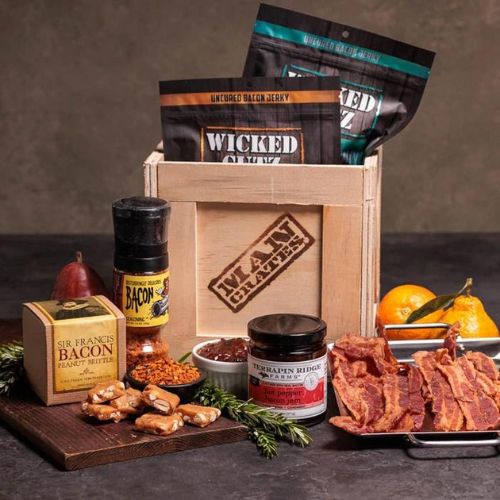 The image shows a wooden gift crate with a black lid and a crowbar attached to it. The crate has a burnt-in Man Crates logo and the words "Baconology Kit" written on it. Inside the crate, there are several items including a bacon press, a bacon grilling rack, a bacon cookbook, and bacon-flavored seasonings. The background is a bright, outdoor setting with green trees and a blue sky. The crate is positioned on a wooden table with a grilling utensil visible in the background.