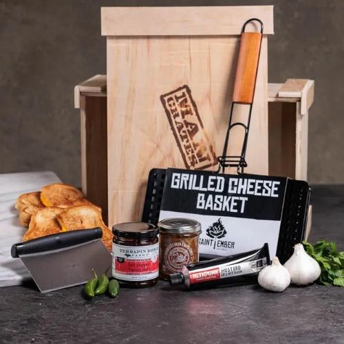 A wooden gift crate with a black lid and a crowbar attached to it. The crate has a burnt-in Man Crates logo and the words "Pizza Grilling Crate" written on it. Inside the crate, there are several items including a pizza stone, a pizza peel, a pizza cutter, and a pizza seasoning kit. The background is a bright, outdoor setting with green trees and a blue sky. The crate is positioned on a wooden table with a grill visible in the background.