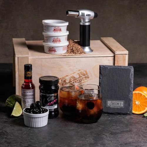 A wooden gift crate with a black lid and a crowbar attached to it. The crate has a burnt-in Man Crates logo and the words "Whiskey Appreciation Crate" written on it. Inside the crate, there are several items including two personalized whiskey glasses, slate coasters, whiskey stones, a wooden crate, and a pocket whiskey journal. The background is a dark, moody setting with a blurred whiskey bottle and glass in the foreground.